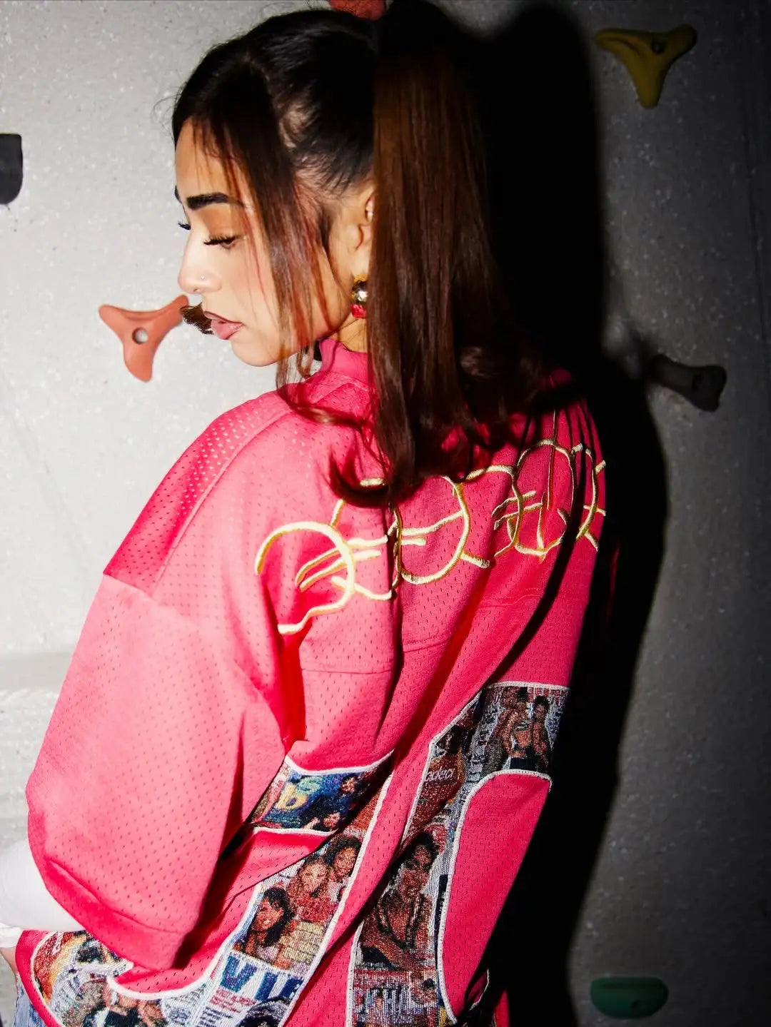 A vibrant pink mesh hoodie featuring gold chain embroidery across the back and a collage of magazine-style images along the lower hem.